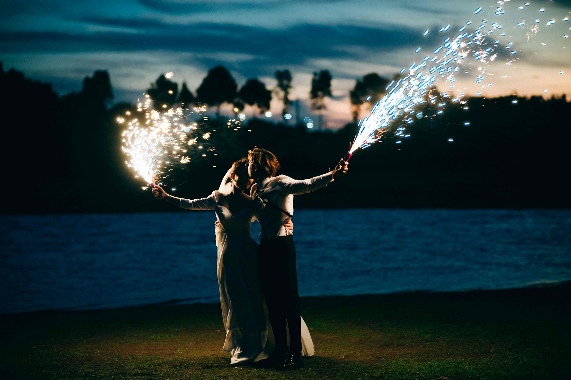 Newlywed couple celebrating with sparklers by a tranquil lake at twilight.