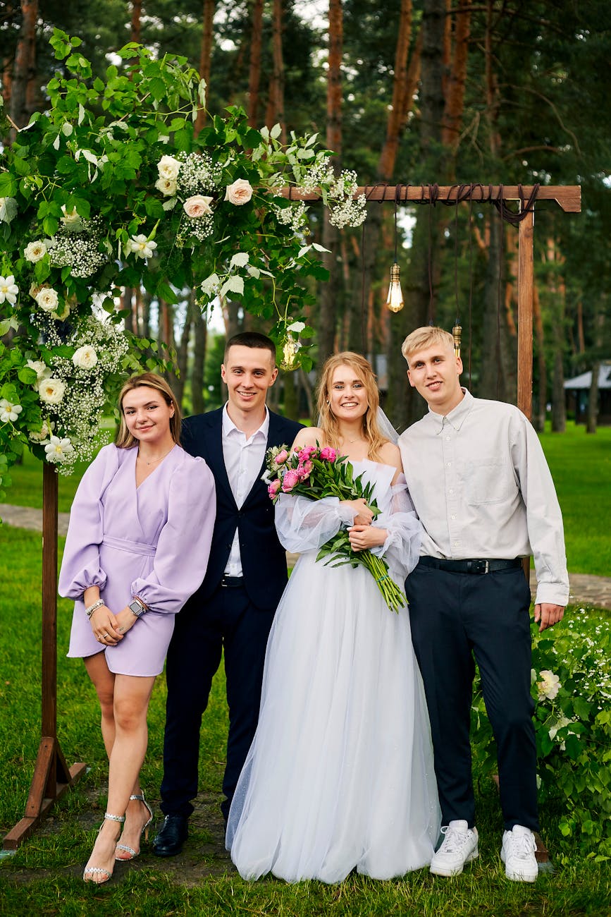 A joyful outdoor wedding photograph featuring the bride, groom, and friends under a floral arch.