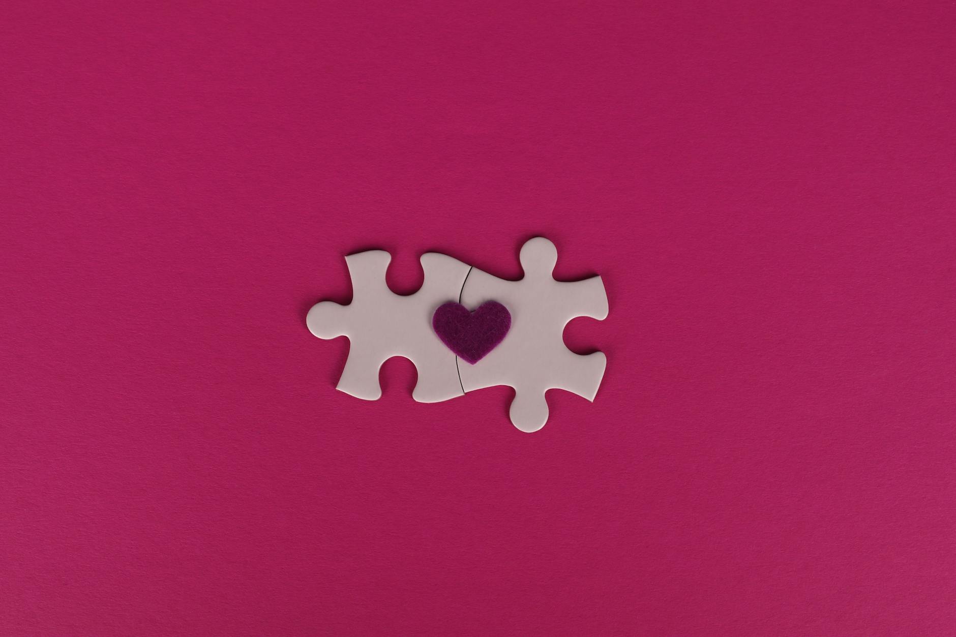 Two white puzzle pieces with a heart symbol on a pink background, symbolizing love and connection.